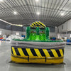 Front view of 46ft Hazardous Commercial Hybrid Inflatable Obstacle Course featuring a yellow and black hazard stripe design. The inflatable structure includes dual green slides, multiple obstacles, and a large yellow and black striped arch. Set up in a spacious warehouse with metal roofing, bright lighting, and various other inflatable products visible in the background.