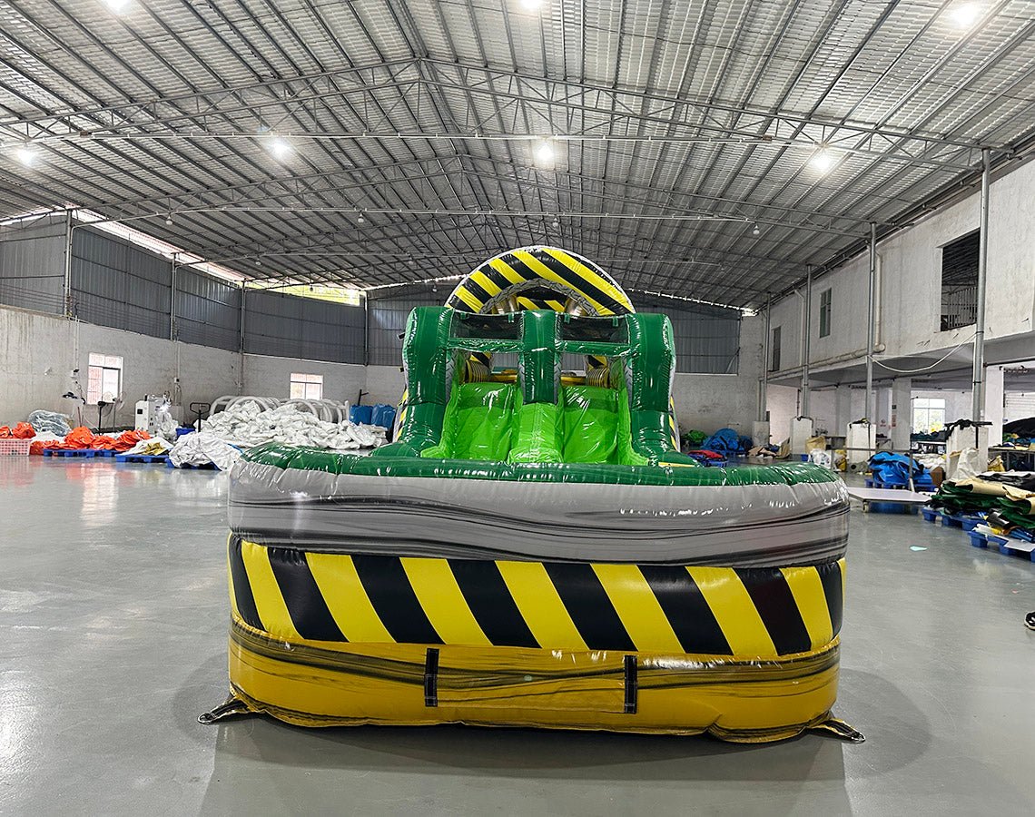 Front view of 46ft Hazardous Commercial Hybrid Inflatable Obstacle Course featuring a yellow and black hazard stripe design. The inflatable structure includes dual green slides, multiple obstacles, and a large yellow and black striped arch. Set up in a spacious warehouse with metal roofing, bright lighting, and various other inflatable products visible in the background.