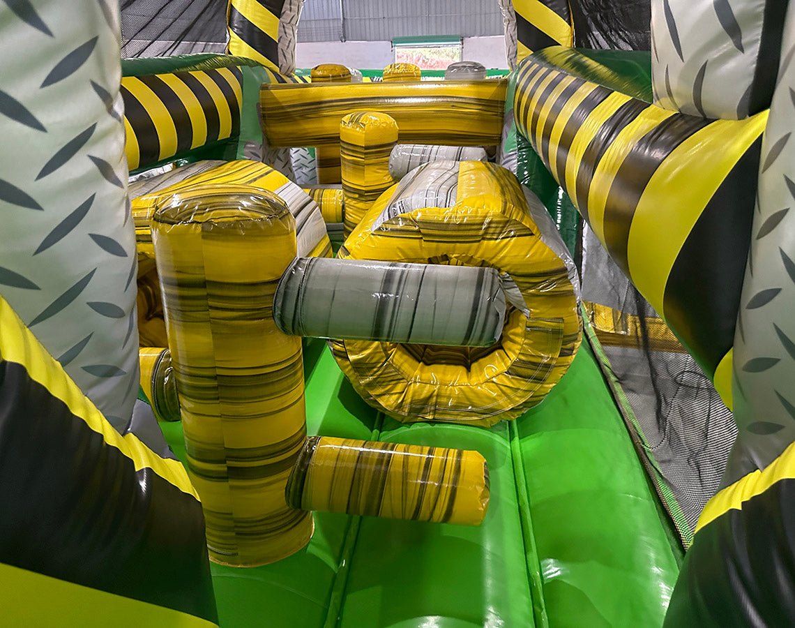 Interior view of 46ft Hazardous Commercial Hybrid Inflatable Obstacle Course showing various obstacles. The image displays yellow inflatable cylinders, a gray inflatable barrel, and yellow and black striped walls. Green flooring is visible, along with mesh netting on the sides for safety.