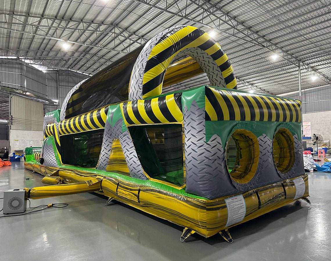 Wide-angle view of 46ft Hazardous Commercial Hybrid Inflatable Obstacle Course set up in a spacious warehouse. The inflatable displays a yellow and black hazard stripe design with multiple obstacles, tunnels, and climbing areas. Other inflatable products and warehouse equipment are visible, showcasing its size and commercial setting.