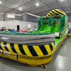 close up front view of 46ft Hazardous Commercial Hybrid Inflatable Obstacle Course showing its full length. The inflatable features a yellow and black hazard stripe theme with green slides, multiple obstacles, and a large arched entrance. Set in a large warehouse with concrete flooring, metal roofing, and other inflatable products visible.