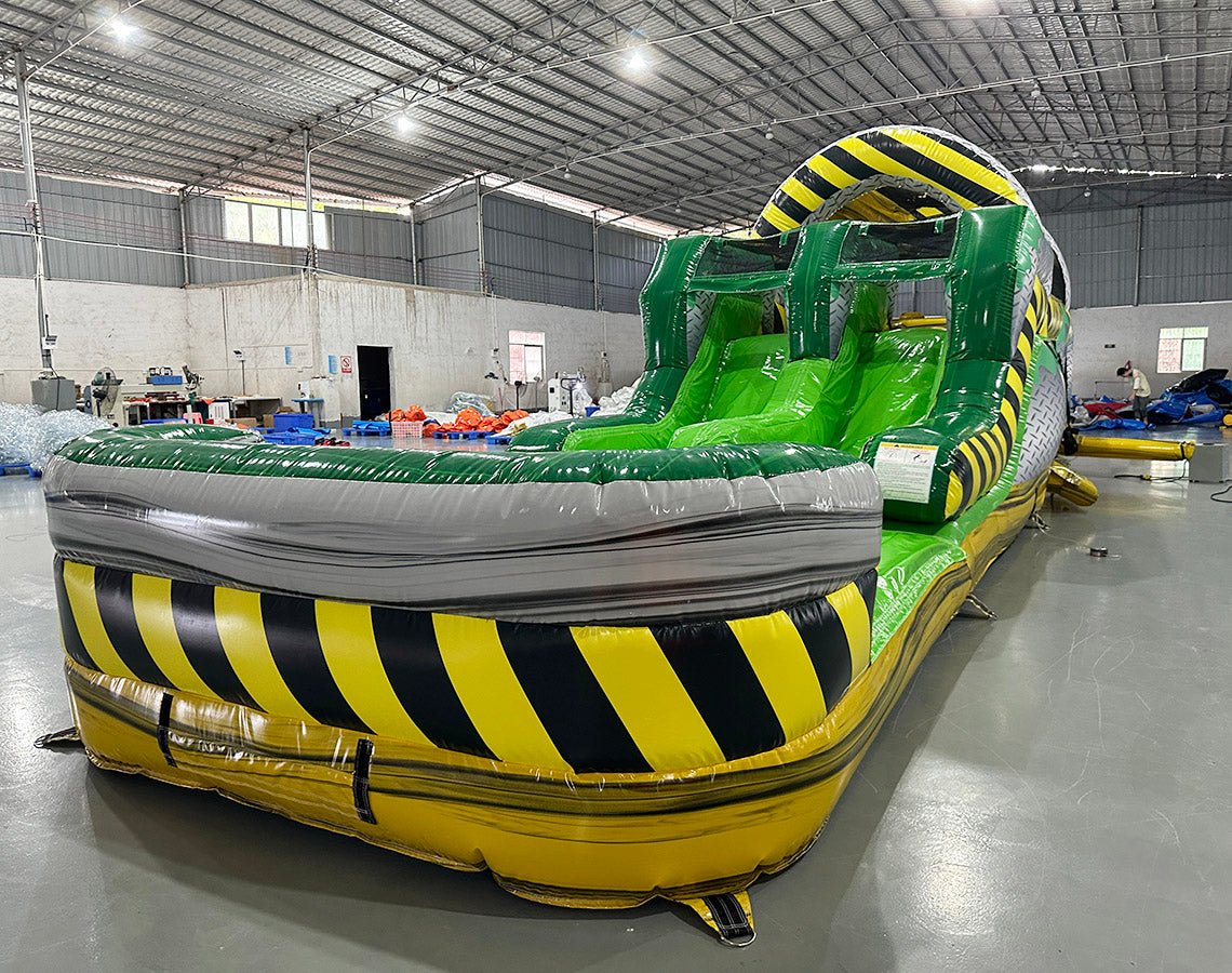 close up front view of 46ft Hazardous Commercial Hybrid Inflatable Obstacle Course showing its full length. The inflatable features a yellow and black hazard stripe theme with green slides, multiple obstacles, and a large arched entrance. Set in a large warehouse with concrete flooring, metal roofing, and other inflatable products visible.