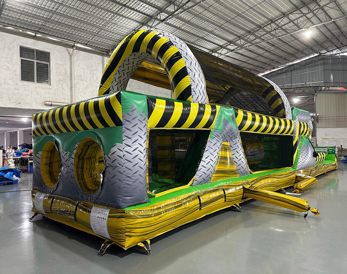 Three-quarter view of 46ft Hazardous Commercial Hybrid Inflatable Obstacle Course in a large warehouse. The structure features yellow and black hazard stripes, circular openings, and various obstacles. The warehouse setting shows metal roofing, support beams, and other inflatable products, demonstrating its commercial application.