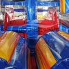 Interior view of 46ft Rip Curl Commercial Hybrid Inflatable Obstacle Course showing dual lanes with red and blue inflatable obstacles. The structure features yellow and blue cylindrical barriers, a central blue divider, and red inflatable platforms. The "BOUNCEWAVE Inflatable Sales" logo is visible on the blue base. Set up in a warehouse with metal roofing visible above.