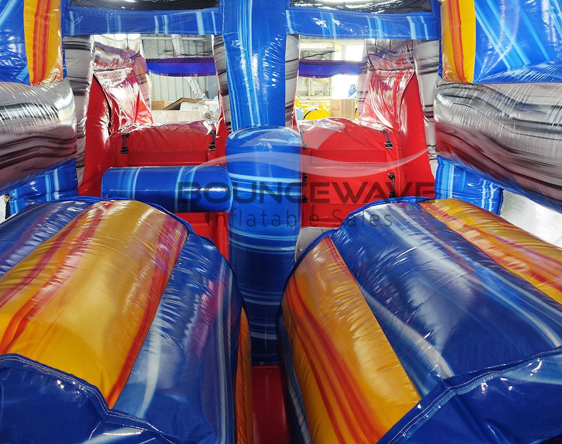 Interior view of 46ft Rip Curl Commercial Hybrid Inflatable Obstacle Course showing dual lanes with red and blue inflatable obstacles. The structure features yellow and blue cylindrical barriers, a central blue divider, and red inflatable platforms. The "BOUNCEWAVE Inflatable Sales" logo is visible on the blue base. Set up in a warehouse with metal roofing visible above.