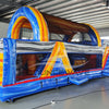 rear side view of 46ft Rip Curl Commercial Hybrid Inflatable Obstacle Course featuring a blue, yellow, and brown color scheme. The inflatable structure includes multiple arches, tunnels, and a red slide. Set up in a large indoor warehouse with metal roofing and various other inflatable products visible in the background.