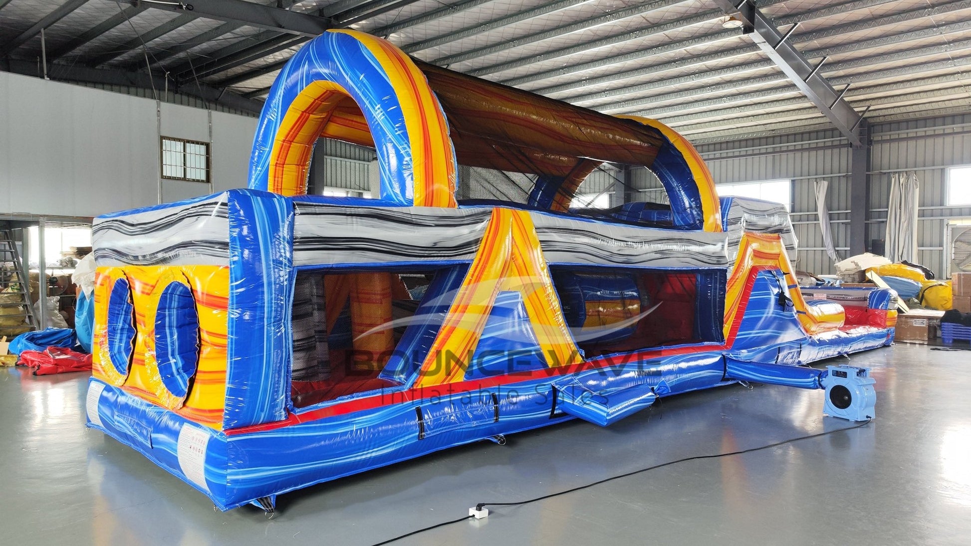 rear side view of 46ft Rip Curl Commercial Hybrid Inflatable Obstacle Course featuring a blue, yellow, and brown color scheme. The inflatable structure includes multiple arches, tunnels, and a red slide. Set up in a large indoor warehouse with metal roofing and various other inflatable products visible in the background.