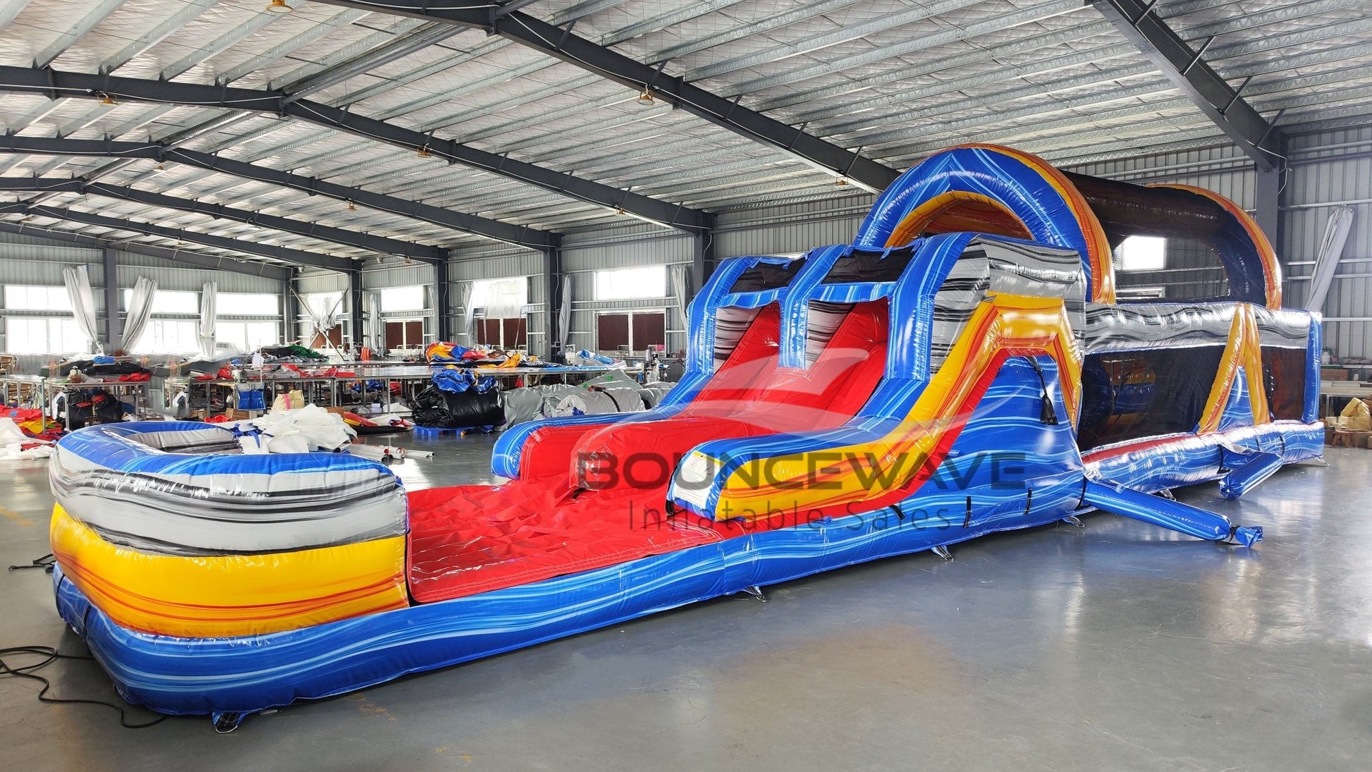 Wide-angle view of 46ft Rip Curl Commercial Hybrid Inflatable Obstacle Course set up in a spacious warehouse. The inflatable displays a blue, yellow, and red color scheme with multiple obstacles, including a large red slide, tunnels, and arches. Various other inflatable products and warehouse equipment are visible, demonstrating its size and commercial setting.