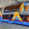 Three-quarter view of 46ft Rip Curl Commercial Hybrid Inflatable Obstacle Course in a large warehouse. The structure features a blue base with yellow and brown accents, multiple arches, tunnels, and a prominent red slide. The warehouse setting shows metal roofing, support beams, and other inflatable products, showcasing its commercial application.