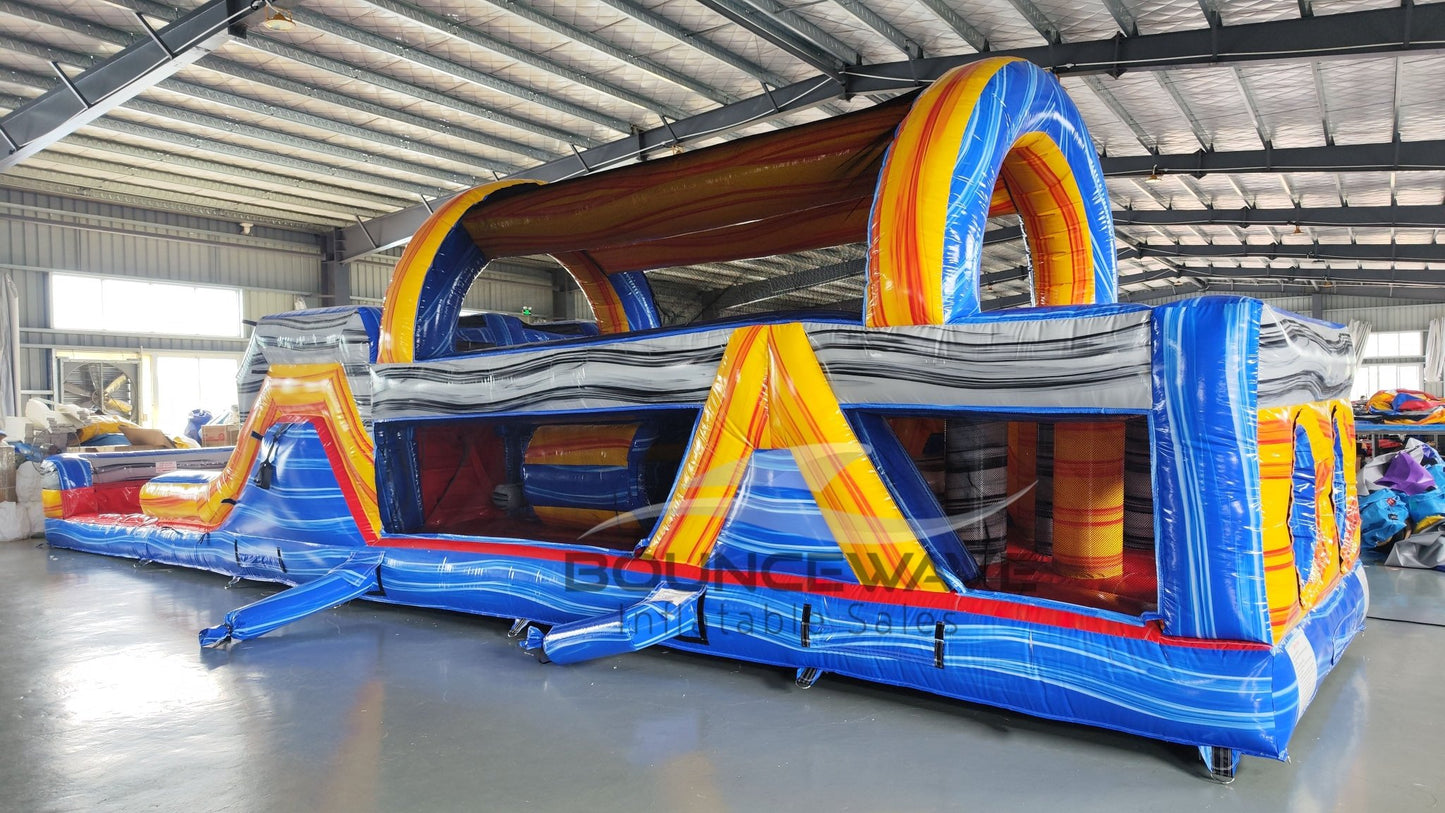 Three-quarter view of 46ft Rip Curl Commercial Hybrid Inflatable Obstacle Course in a large warehouse. The structure features a blue base with yellow and brown accents, multiple arches, tunnels, and a prominent red slide. The warehouse setting shows metal roofing, support beams, and other inflatable products, showcasing its commercial application.