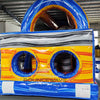 Rear view of 46ft Rip Curl Commercial Hybrid Inflatable Obstacle Course set up in a large indoor warehouse. The inflatable features a blue base with yellow accents, dual circular entrances, and a large blue and yellow arch on top. A gray wavy pattern decorates the upper section. Safety instructions are visible on the base. Various other inflatable products and warehouse equipment are visible in the background.