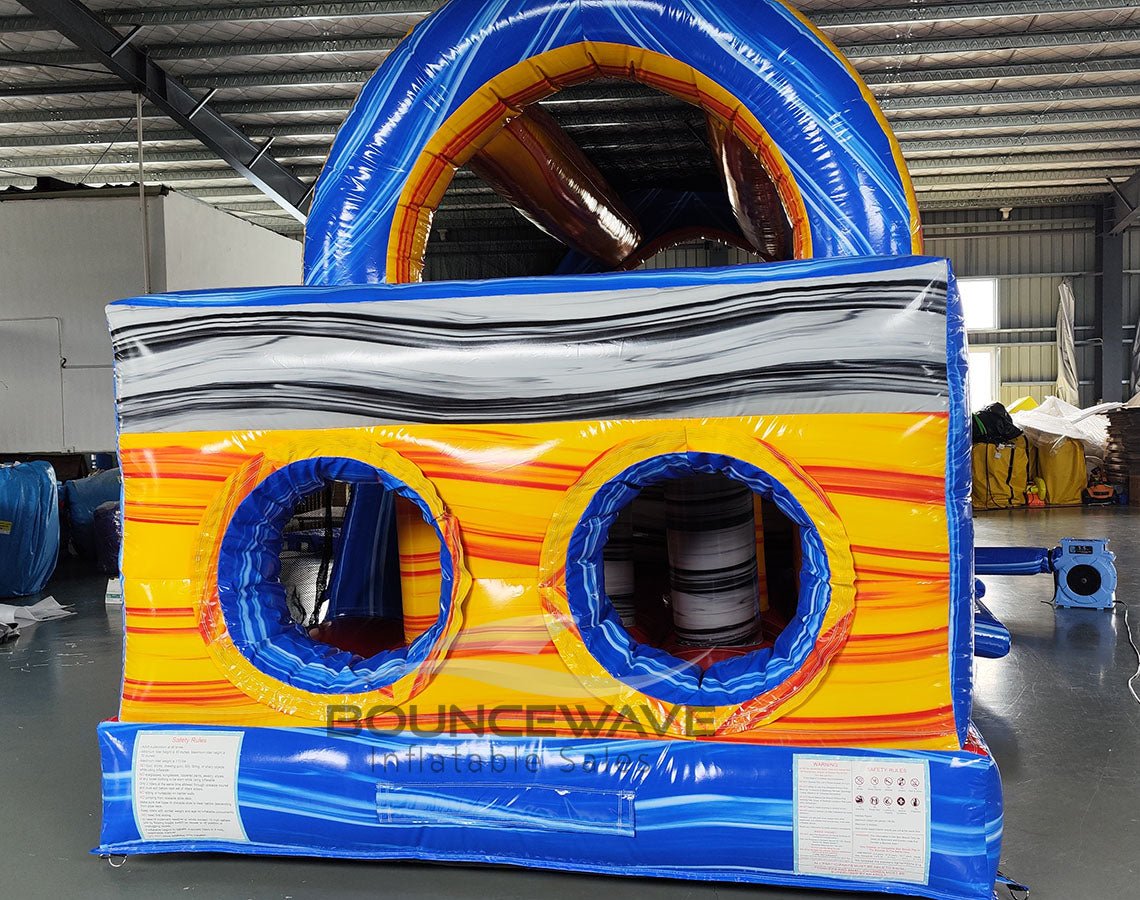 Rear view of 46ft Rip Curl Commercial Hybrid Inflatable Obstacle Course set up in a large indoor warehouse. The inflatable features a blue base with yellow accents, dual circular entrances, and a large blue and yellow arch on top. A gray wavy pattern decorates the upper section. Safety instructions are visible on the base. Various other inflatable products and warehouse equipment are visible in the background.