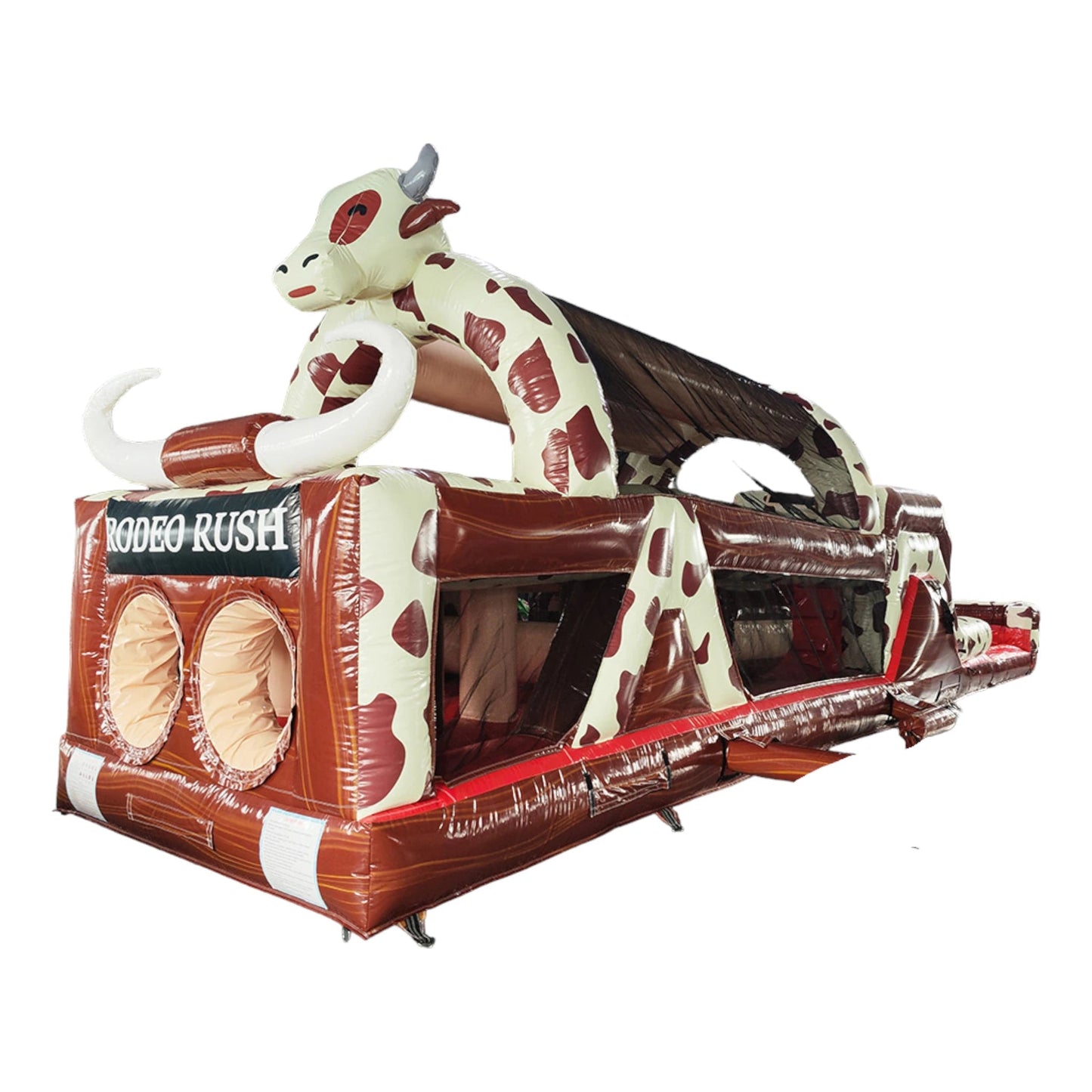 Side view of 46ft Rodeo Rush Commercial Hybrid Inflatable Obstacle Course featuring a cow-print design. The structure includes a large inflatable cow figure on top, dual entrance tunnels, and a brown base with "RODEO RUSH" text. Set up in an indoor warehouse with metal roofing and various equipment visible in the background.