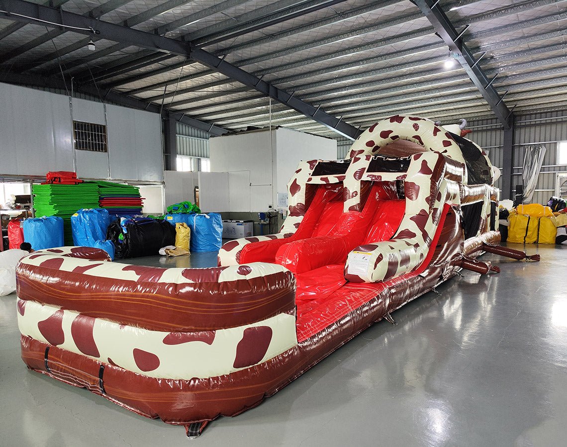 Side view of 46ft Rodeo Rush Commercial Hybrid Inflatable Obstacle Course showing its full length. The inflatable features a cow-print design with dual red slides emerging from the structure. A large inflatable cow figure arches over the top. Various inflatable obstacles and colorful storage bags are visible in the warehouse setting.