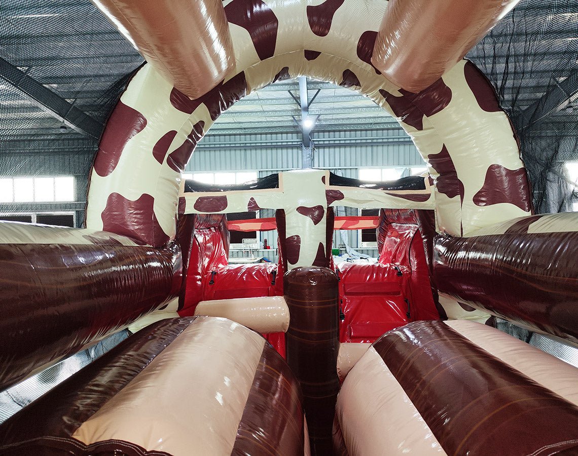 Interior view of 46ft Rodeo Rush Commercial Hybrid Inflatable Obstacle Course showing dual lanes with red inflatable obstacles. The structure features a cow-print design on the walls and ceiling, with a central brown divider. Red inflatable cylinders and platforms are visible as part of the obstacle course.