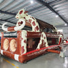 Front view of 46ft Rodeo Rush Commercial Hybrid Inflatable Obstacle Course featuring a large inflatable cow figure arching over the structure. The inflatable has a brown base with "RODEO RUSH" text, dual circular entrances, and a cow-print design. Set up in a large indoor warehouse with metal roofing and various other inflatable products visible in the background.