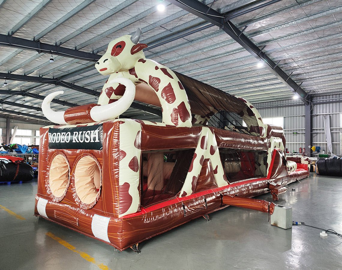 Front view of 46ft Rodeo Rush Commercial Hybrid Inflatable Obstacle Course featuring a large inflatable cow figure arching over the structure. The inflatable has a brown base with "RODEO RUSH" text, dual circular entrances, and a cow-print design. Set up in a large indoor warehouse with metal roofing and various other inflatable products visible in the background.