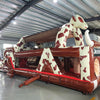 Wide-angle view of 46ft Rodeo Rush Commercial Hybrid Inflatable Obstacle Course in a large warehouse. The inflatable displays a cow-print design with a large cow figure arching over the structure, dual slides, and multiple obstacles. Various other inflatable products and warehouse equipment are visible, showcasing its size and commercial setting.