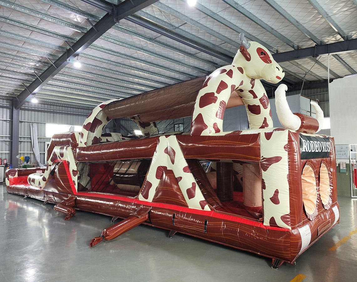 Wide-angle view of 46ft Rodeo Rush Commercial Hybrid Inflatable Obstacle Course in a large warehouse. The inflatable displays a cow-print design with a large cow figure arching over the structure, dual slides, and multiple obstacles. Various other inflatable products and warehouse equipment are visible, showcasing its size and commercial setting.