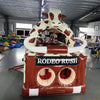 Close-up front view of 46ft Rodeo Rush Commercial Hybrid Inflatable Obstacle Course focusing on the entrance area. Features a large inflatable cow figure, "RODEO RUSH" text on a brown base, and dual circular entrances. Set in a warehouse with various inflatable products and equipment visible in the background.