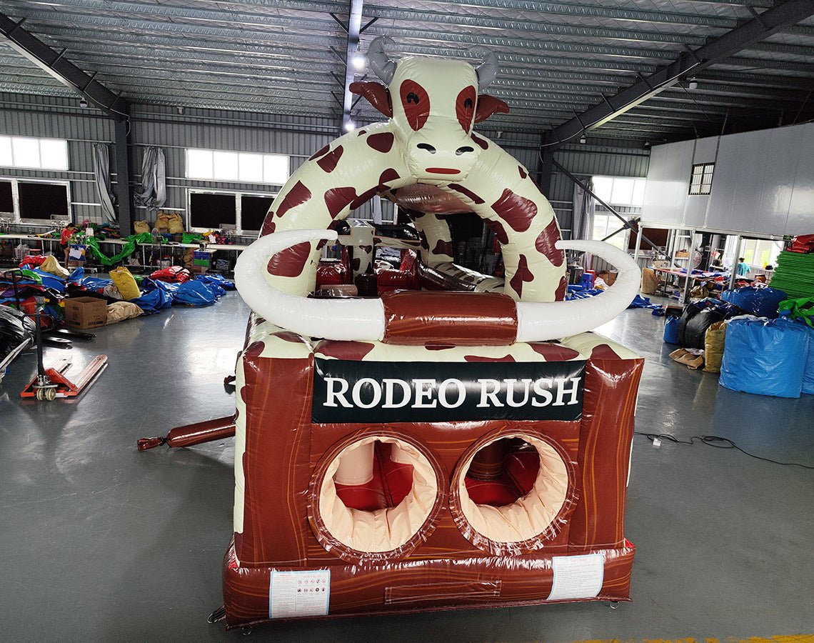 Close-up front view of 46ft Rodeo Rush Commercial Hybrid Inflatable Obstacle Course focusing on the entrance area. Features a large inflatable cow figure, "RODEO RUSH" text on a brown base, and dual circular entrances. Set in a warehouse with various inflatable products and equipment visible in the background.