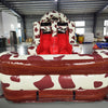 Front view of 46ft Rodeo Rush Commercial Hybrid Inflatable Obstacle Course showing dual red slides emerging from a cow-print structure. The inflatable features a large cow figure on top and a brown base with circular entrances. Set in a spacious warehouse with concrete flooring and other inflatable products visible.