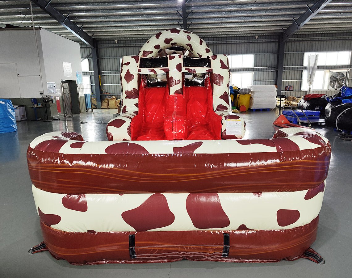 Front view of 46ft Rodeo Rush Commercial Hybrid Inflatable Obstacle Course showing dual red slides emerging from a cow-print structure. The inflatable features a large cow figure on top and a brown base with circular entrances. Set in a spacious warehouse with concrete flooring and other inflatable products visible.