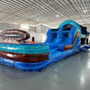 Front view of 46ft South Beach Commercial Hybrid Inflatable Obstacle Course featuring a tropical theme. The structure has dual blue circular entrances, brown inflatable palm tree trunks with turquoise leaves, and large sunglasses on top displaying a beach scene. Set up in a large indoor warehouse with metal roofing, concrete flooring, and various other inflatable products visible in the background.