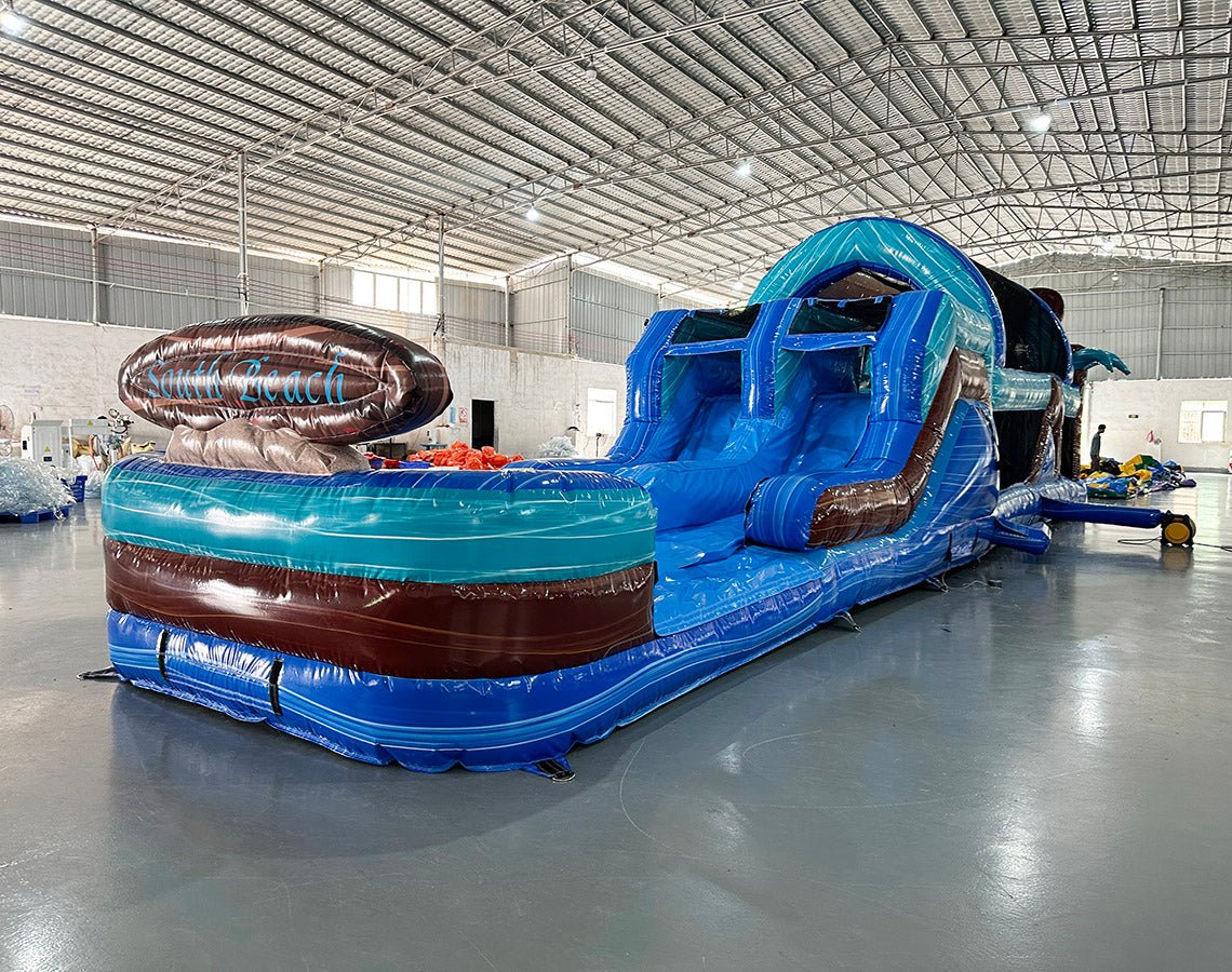 Front view of 46ft South Beach Commercial Hybrid Inflatable Obstacle Course featuring a tropical theme. The structure has dual blue circular entrances, brown inflatable palm tree trunks with turquoise leaves, and large sunglasses on top displaying a beach scene. Set up in a large indoor warehouse with metal roofing, concrete flooring, and various other inflatable products visible in the background.