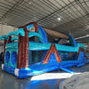 Three-quarter view of 46ft South Beach Commercial Hybrid Inflatable Obstacle Course in a spacious warehouse. The inflatable displays a tropical theme with brown palm tree trunks, turquoise leaves, and large sunglasses on top. Multiple obstacles, tunnels, and a long blue base are visible. Other inflatable products and warehouse materials are visible in the background.