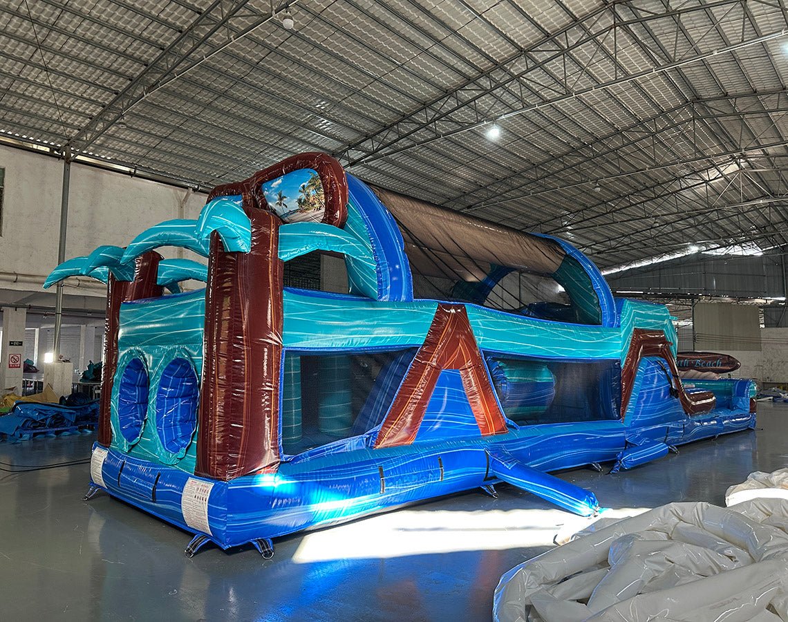 Three-quarter view of 46ft South Beach Commercial Hybrid Inflatable Obstacle Course in a spacious warehouse. The inflatable displays a tropical theme with brown palm tree trunks, turquoise leaves, and large sunglasses on top. Multiple obstacles, tunnels, and a long blue base are visible. Other inflatable products and warehouse materials are visible in the background.