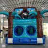 Front view of 46ft South Beach Commercial Hybrid Inflatable Obstacle Course set up in a large indoor warehouse. The inflatable features brown palm tree trunks, turquoise palm leaves, and large sunglasses with a beach scene. Dual blue circular entrances are visible at the base. Metal roofing and various other inflatable products are visible in the background.