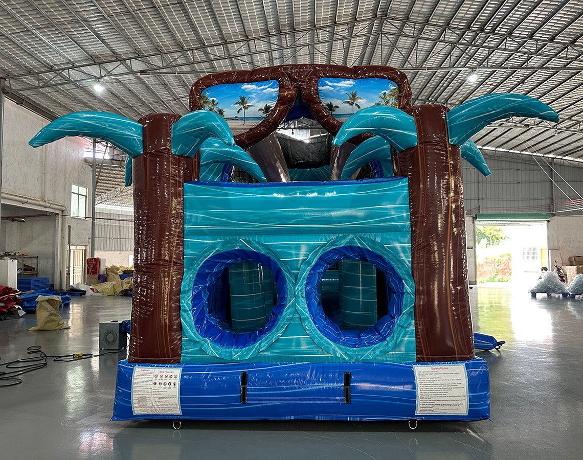 Front view of 46ft South Beach Commercial Hybrid Inflatable Obstacle Course set up in a large indoor warehouse. The inflatable features brown palm tree trunks, turquoise palm leaves, and large sunglasses with a beach scene. Dual blue circular entrances are visible at the base. Metal roofing and various other inflatable products are visible in the background.