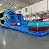 Side view of 46ft South Beach Commercial Hybrid Inflatable Obstacle Course showing its full length. The inflatable features a blue base, brown palm tree trunks with turquoise leaves, and large sunglasses on top. Multiple obstacles and tunnels are visible along its length. Set in a spacious warehouse with metal roofing, bright lighting, and other inflatable products in the background.