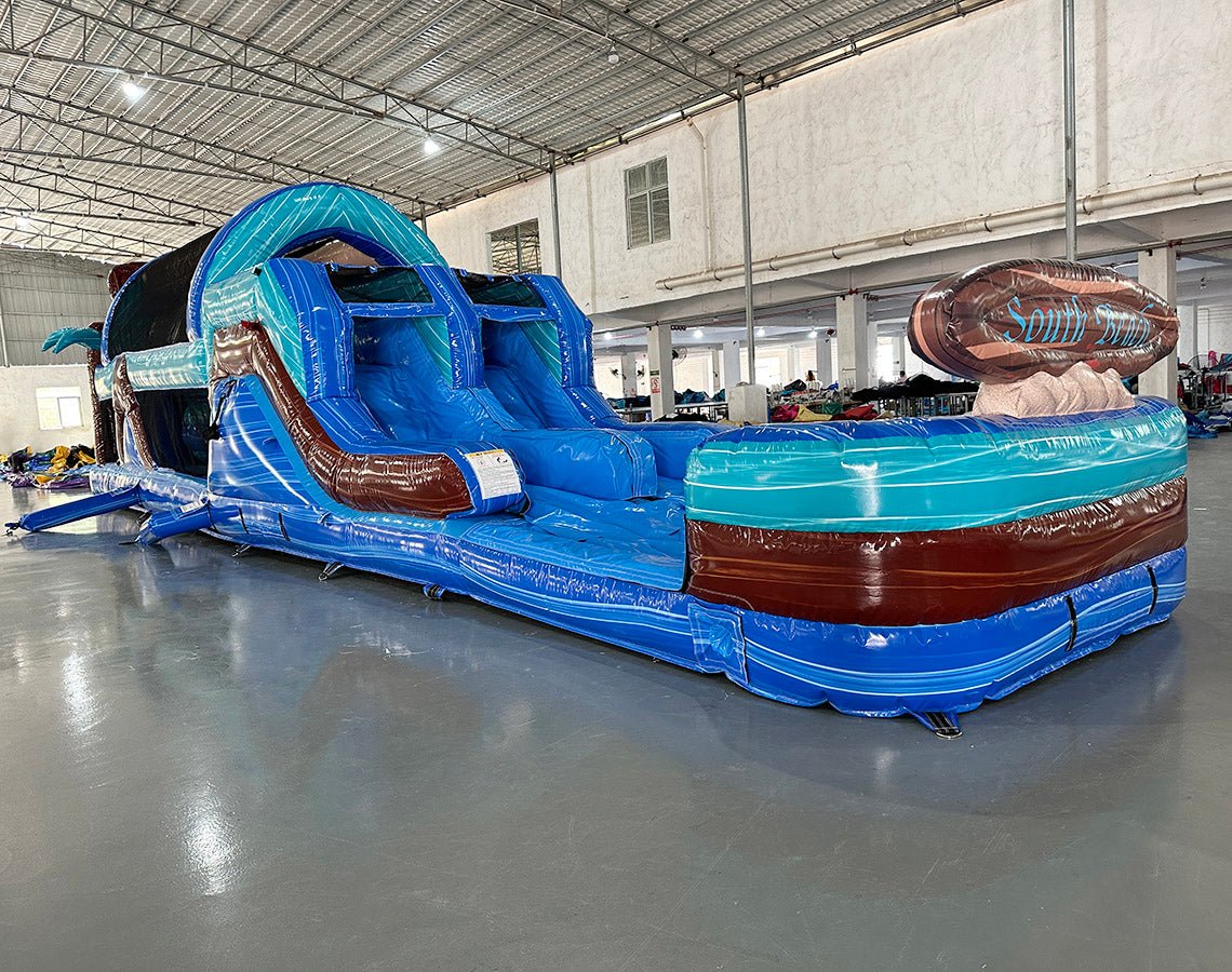 Side view of 46ft South Beach Commercial Hybrid Inflatable Obstacle Course showing its full length. The inflatable features a blue base, brown palm tree trunks with turquoise leaves, and large sunglasses on top. Multiple obstacles and tunnels are visible along its length. Set in a spacious warehouse with metal roofing, bright lighting, and other inflatable products in the background.