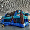 Wide-angle view of 46ft South Beach Commercial Hybrid Inflatable Obstacle Course showing its full length. The structure features brown palm tree trunks, turquoise leaves, large sunglasses on top, and a long blue base with multiple obstacles. Set up in a warehouse with metal roofing and bright lighting, showcasing its scale and commercial application.