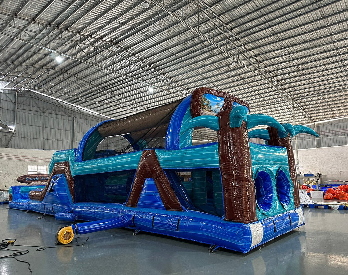 Wide-angle view of 46ft South Beach Commercial Hybrid Inflatable Obstacle Course showing its full length. The structure features brown palm tree trunks, turquoise leaves, large sunglasses on top, and a long blue base with multiple obstacles. Set up in a warehouse with metal roofing and bright lighting, showcasing its scale and commercial application.