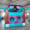 Rear view of 53ft XL Island Flow Commercial Hybrid Inflatable Obstacle Course featuring a tropical theme. The structure has dual blue slides, inflatable palm trees with brown trunks and turquoise leaves, and a colorful base with turquoise, pink, and yellow sections. A large inflatable sunglasses with a beach scene tops the entrance. Set up in a spacious warehouse with metal roofing and various other inflatable products visible in the background.