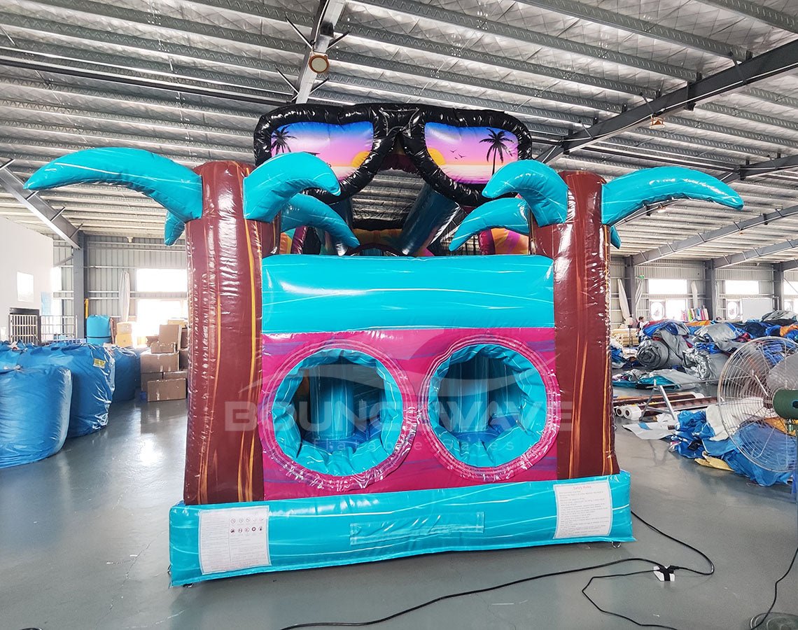 Rear view of 53ft XL Island Flow Commercial Hybrid Inflatable Obstacle Course featuring a tropical theme. The structure has dual blue slides, inflatable palm trees with brown trunks and turquoise leaves, and a colorful base with turquoise, pink, and yellow sections. A large inflatable sunglasses with a beach scene tops the entrance. Set up in a spacious warehouse with metal roofing and various other inflatable products visible in the background.