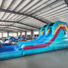 Wide-angle view of 53ft XL Island Flow Commercial Hybrid Inflatable Obstacle Course set up in a large indoor warehouse. The inflatable features dual blue slides, inflatable palm trees, and a long turquoise base with colorful accents. A large inflatable sunglasses with a tropical scene sits atop the structure. Various other inflatable products and storage containers are visible in the foreground and background.