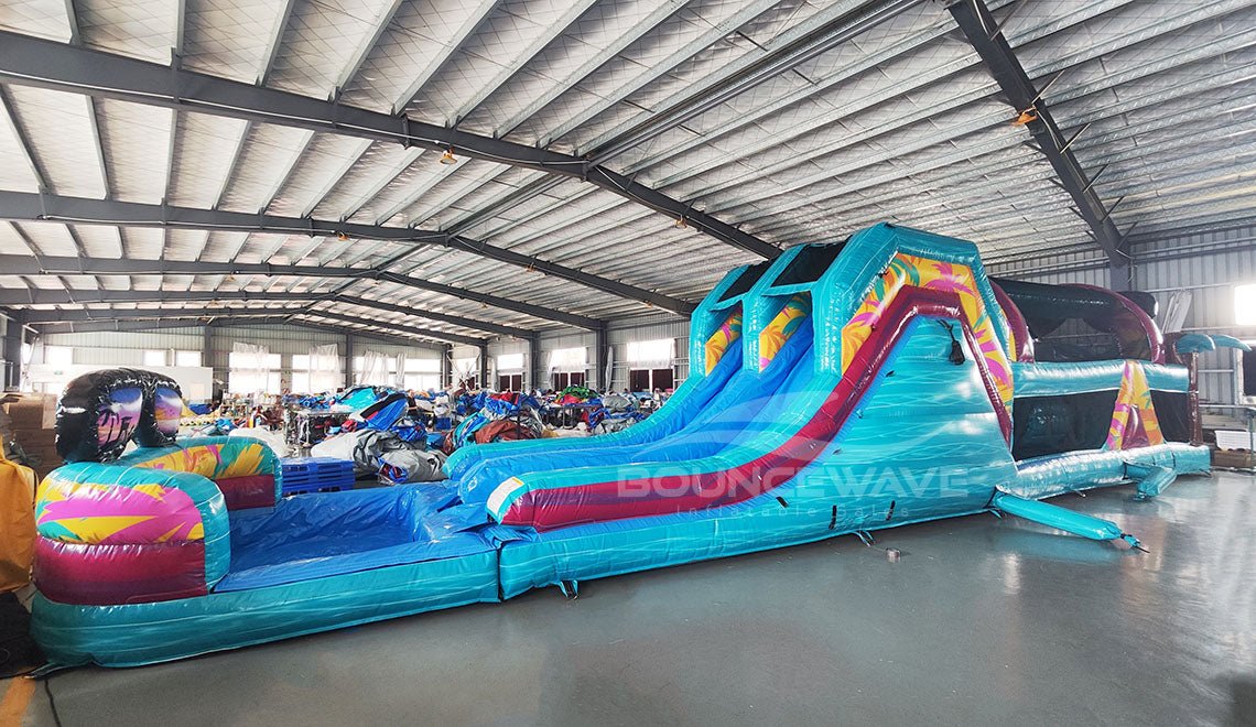 Wide-angle view of 53ft XL Island Flow Commercial Hybrid Inflatable Obstacle Course set up in a large indoor warehouse. The inflatable features dual blue slides, inflatable palm trees, and a long turquoise base with colorful accents. A large inflatable sunglasses with a tropical scene sits atop the structure. Various other inflatable products and storage containers are visible in the foreground and background.