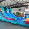 Front view of 53ft XL Island Flow Commercial Hybrid Inflatable Obstacle Course featuring a tropical theme. The structure has dual blue slides, inflatable palm trees with brown trunks and turquoise leaves, and a colorful base with turquoise, pink, and yellow sections. A large inflatable sunglasses with a beach scene tops the entrance. Set up in a spacious warehouse with metal roofing and various other inflatable products visible in the background.