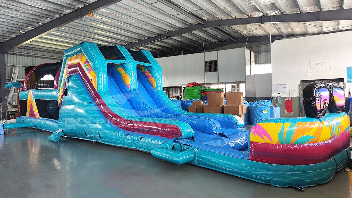 Front view of 53ft XL Island Flow Commercial Hybrid Inflatable Obstacle Course featuring a tropical theme. The structure has dual blue slides, inflatable palm trees with brown trunks and turquoise leaves, and a colorful base with turquoise, pink, and yellow sections. A large inflatable sunglasses with a beach scene tops the entrance. Set up in a spacious warehouse with metal roofing and various other inflatable products visible in the background.