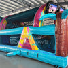 Side view of 53ft XL Island Flow Commercial Hybrid Inflatable Obstacle Course showing its full length. The inflatable features a turquoise base, dual blue slides, brown palm tree trunks with turquoise leaves, and a large inflatable sunglasses on top. Colorful tropical-themed panels decorate the sides. Set in a large indoor warehouse with metal roofing, concrete flooring, and various other inflatable products and equipment visible in the background.