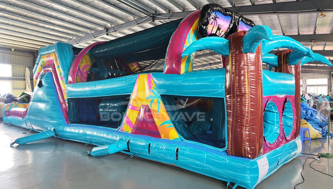 Side view of 53ft XL Island Flow Commercial Hybrid Inflatable Obstacle Course showing its full length. The inflatable features a turquoise base, dual blue slides, brown palm tree trunks with turquoise leaves, and a large inflatable sunglasses on top. Colorful tropical-themed panels decorate the sides. Set in a large indoor warehouse with metal roofing, concrete flooring, and various other inflatable products and equipment visible in the background.