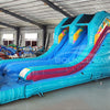 Front view of 53ft XL Island Flow Commercial Hybrid Inflatable Obstacle Course in a warehouse setting. The structure features dual blue slides, inflatable palm trees, and a turquoise base with colorful accents. Large sunglasses with a beach scene sit atop the entrance. The warehouse interior shows metal roofing, support beams, and various other inflatable products, demonstrating its size and commercial application.