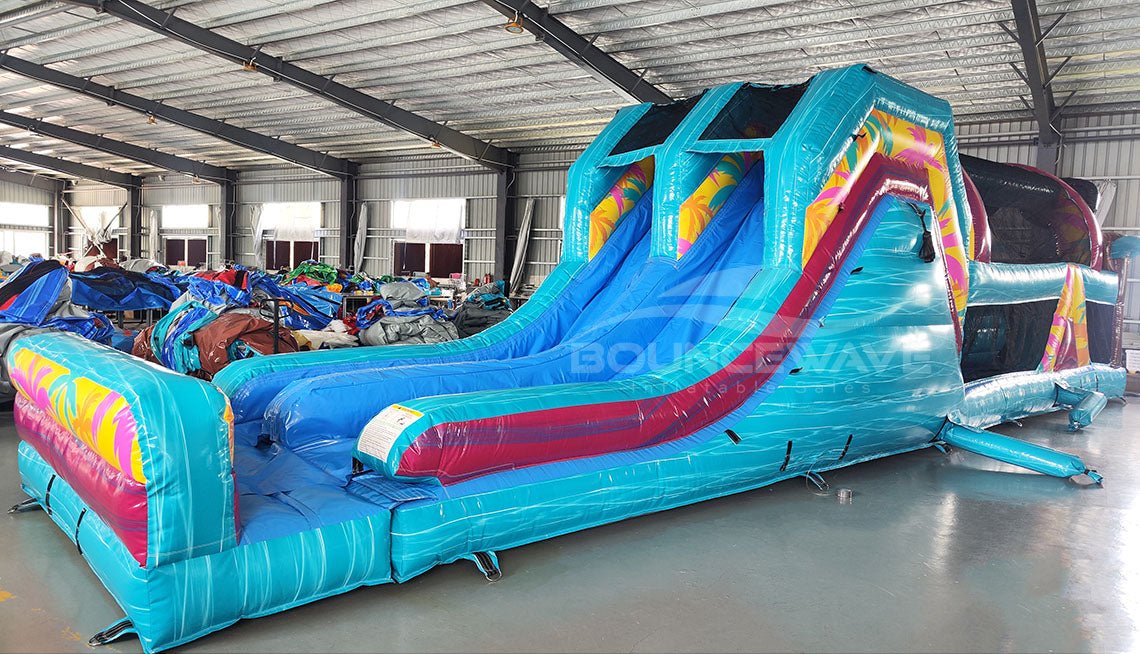 Front view of 53ft XL Island Flow Commercial Hybrid Inflatable Obstacle Course in a warehouse setting. The structure features dual blue slides, inflatable palm trees, and a turquoise base with colorful accents. Large sunglasses with a beach scene sit atop the entrance. The warehouse interior shows metal roofing, support beams, and various other inflatable products, demonstrating its size and commercial application.