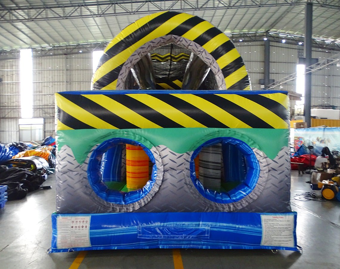 Rear view of 53ft XL Radioactive Run Commercial Hybrid Inflatable Obstacle Course showing the entrance area. Features a large yellow and black striped arch, dual circular entrances with blue trim, and a diamond plate pattern base. Safety instructions are visible on the front panel. Set in a warehouse with concrete flooring and various other inflatable products visible in the background.