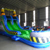 Front angel view of 53ft XL Radioactive Run Commercial Hybrid Inflatable Obstacle Course featuring a hazardous materials theme. The structure has yellow and black striped sections, a diamond plate pattern base, and dual green slides. Set up in a large indoor warehouse with metal roofing and various other inflatable products visible in the background.