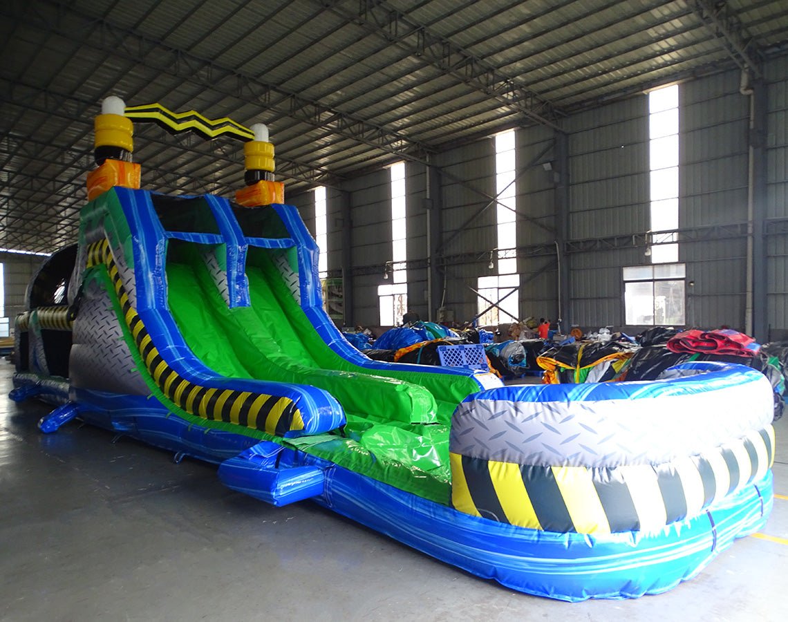 Front angel view of 53ft XL Radioactive Run Commercial Hybrid Inflatable Obstacle Course featuring a hazardous materials theme. The structure has yellow and black striped sections, a diamond plate pattern base, and dual green slides. Set up in a large indoor warehouse with metal roofing and various other inflatable products visible in the background.